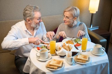 Gülümseyen yetişkin beyaz bir çift kahvaltı yapıyor ve sabahları birbirlerine bakıyorlar. Dinlenme, tatil ve seyahat kavramı. İlişki fikri