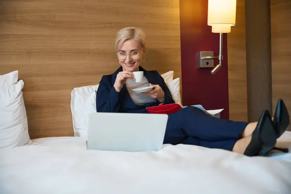 Smiling adult caucasian businesswoman drinking tea or coffee from cup and watching laptop on bed in hotel room at daytime. Concept of business trip, vacation and travelling