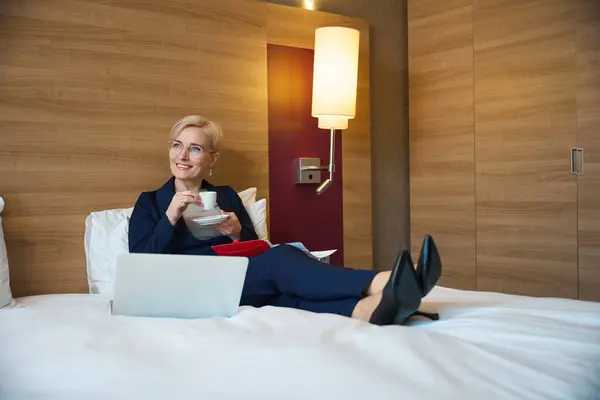 Smiling adult caucasian businesswoman drinking tea or coffee from cup and looking away on bed in hotel room at daytime. Concept of business trip, vacation and travelling