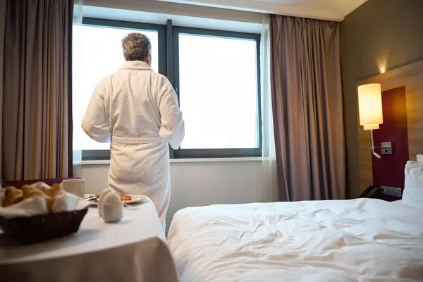 Back view of man wearing bathrobe standing and looking in window having breakfast in hotel room at morning. Concept of rest, vacation and travelling
