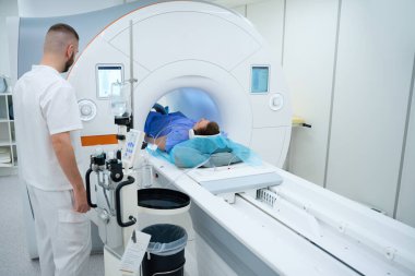 Kulaklıklı hasta tarama masasında yatarken radyograf MRI gantry kontrol panelinde düğmeye basıyor ve bacağı sarmalanıyor.
