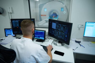 Kontrol odasındaki masada oturan MR teknisyeni bilgisayar monitöründeki kafa MRI taramalarını izliyor.