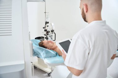 Elinde tablet bilgisayarla Radyolojik Teknoloji uzmanı kontrastlı enjektörün yanındaki bilgisayarlı tomografi masasında yatan adamın önünde duruyor.