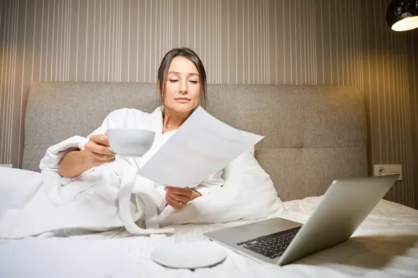 Young focused pretty caucasian businesswoman wearing bathrobe with cup of tea or coffee watching document on bed in hotel room at morning time. Concept of business trip, vacation and travelling