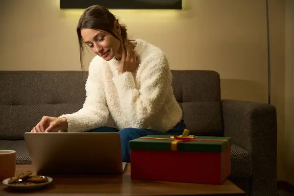 圣诞节或新年期间 年轻而漂亮的 专心致志的高加索女人坐在沙发上 晚上在家里带着礼品盒 一边看笔记本电脑 庆祝寒假的概念 — 图库照片