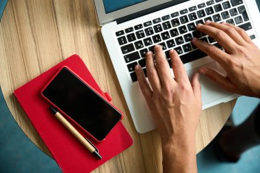 Erkek eli klavyede bir şeyler yazarken dizüstü bilgisayarın, akıllı telefonun, defterin ve kalemin en üstteki görüntüsü.