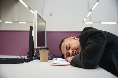 Genç Afrika kökenli Amerikalı bir çalışan bilgisayarın yanındaki çalışma yerinde uyurken masada yatıyor.