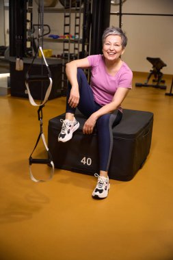 Spor kıyafetleri giyen beyaz kadın spor salonunda oturuyor ve spor salonunda molada kameraya bakıyor. Modern sağlıklı yaşam tarzı kavramı