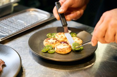 Lokantadaki masada peynirli kekler için krep spatulası ve cımbızla birlikte kivi dilimleri koyan kırpık erkek şef. Lezzetli sağlıklı yemek kavramı