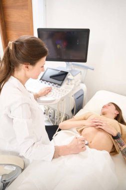 Yetişkin kadın jinekoloğun klinikte genç bir kadının karnının ultrason taramasını yaparken yüksek açılı görüntüsü. Hamilelik ve annelik kavramı