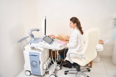 Kafkasyalı kadın jinekoloğun klinikteki tıbbi kanepede hamile Avrupalı bir kadının karnının ultrason taramasını yaparken yan görüntüsü. Hamilelik ve annelik kavramı