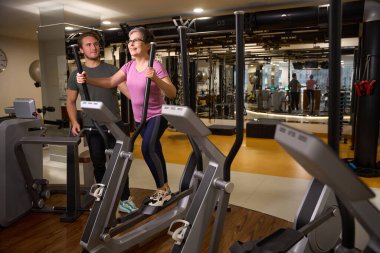Genç erkek antrenör spor giyimli orta yaşlı beyaz bir kadına yardım ediyor spor salonunda koşu bandında koşuyor. Modern sağlıklı yaşam tarzı kavramı