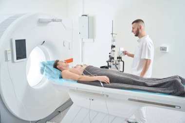 Radyolojik teknoloji kontrastlı enjektörü çalıştırırken ECG elektrotları olan hastayı tomografi masasında sırtüstü yatarken izliyor.