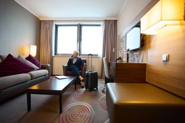 Yetişkin beyaz iş kadını gündüzleri otel odasındaki koltukta akıllı telefondan konuşuyor. İş gezisi, tatil ve seyahat kavramı