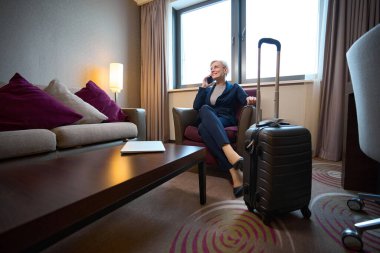 Gündüz vakti otel odasındaki koltukta akıllı telefondan konuşan gülümseyen beyaz iş kadını. İş gezisi, tatil ve seyahat kavramı