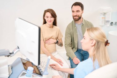 Beyaz kadın jinekolog ve Avrupalı genç bir çift klinikte hamile kadının karnının ultrasonla taranmasını izliyorlar. Hamilelik ve babalık kavramı