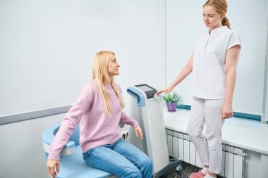 Belden aşağısı ile ilgili sorunları olan genç bir kadın elektromanyetik uyarılma koltuğunda, uzman hemşire gözetiminde, terapi prosedürüne hazırlanıyor.