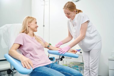 Yüksek nitelikli tıp laboratuarı hemşiresi, sıcak muayene koltuğunda, sağlık kontrolünde oturan bir kadın hastadan kan örneği alacak.