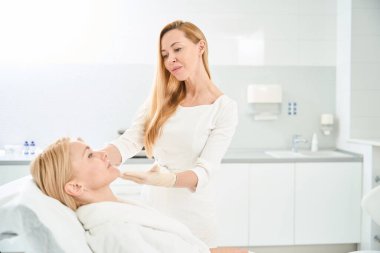 Biyolojik canlandırma prosedüründen sonra müşterinin yüzüne bakan çekici olgun kozmetikçi, ilk sonuçlardan, kişiselleştirilmiş tedaviden, estetik tıp kliniğinden memnun.