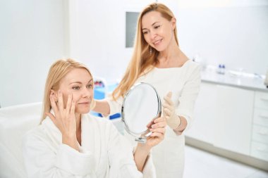 Gülümseyen kadın estetik tıp kliniğinin müşterisi yara izinden ve gözlerinin etrafındaki derinin kaldırılmasından memnun, aynadaki yansımasına bakıyor, iyi servis.