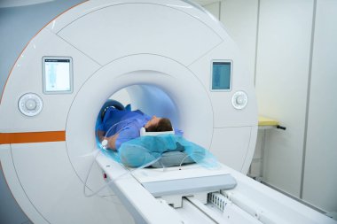 Gürültü önleyici kulaklıklar takan erkek ve dizlerinin etrafına sarılmış bobin MRI makinesinin içinde, masanın üzerinde yatıyor.