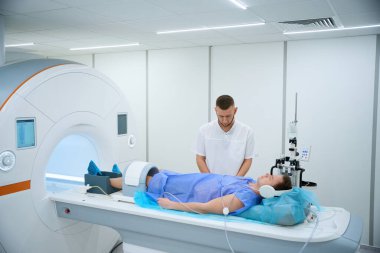 Radiographer standing near patient wearing noise-canceling headphones and knee coil positioned on MRI table clipart