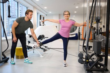 Erkek antrenör, spor kıyafetleri giyen orta yaşlı beyaz bir kadının spor salonunda bacak egzersizi yapmasına yardım ediyor. Modern sağlıklı yaşam tarzı kavramı