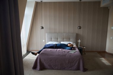 Yorgun Afrika kökenli Amerikalı erkek girişimcinin gündüz vakti otel odasında dizüstü bilgisayarla yatıp dinlenmesi. İş gezisi, tatil ve seyahat kavramı