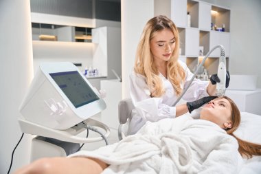Bulanık güzellik salonunda genç beyaz bir kadının IPL fotorejuvenation cihazıyla kafa derisinin mikronedle kaldırılmasını yapan Avrupalı kadın güzellik uzmanı. Yüz bakımı ve gençleştirme kavramı