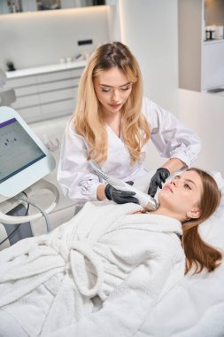 Bulanık güzellik salonunda genç beyaz bir kadının IPL fotorejuvenation cihazıyla boyun derisinin mikronedle kaldırılmasını yapan Avrupalı kadın güzellik uzmanı. Vücut bakımı ve gençleştirme kavramı