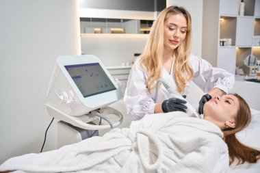 Bulanık güzellik salonunda genç beyaz bir kadının IPL fotorejuvenation cihazıyla boyun derisinin mikronedle kaldırılmasını yapan Avrupalı kadın kozmetik uzmanı. Vücut bakımı ve gençleştirme kavramı