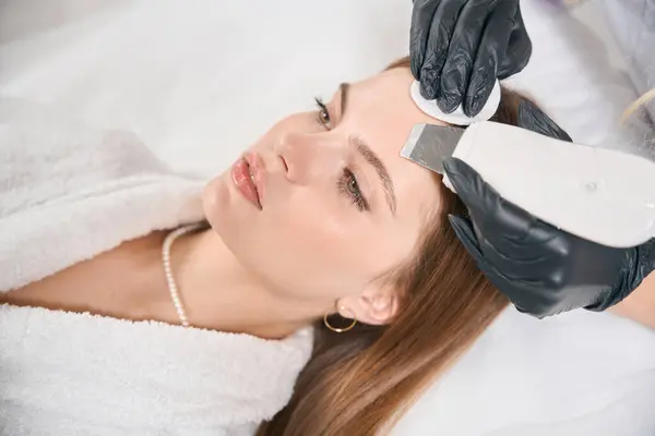 stock image Partial female cosmetologist doing ultrasonic cleaning of face skin with IPL photorejuvenation device of young caucasian woman in blurred beauty salon. Concept of face skin care and rejuvenation