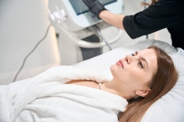 Kısmen bulanık kadın güzellik uzmanı güzellik salonundaki kanepede bornoz giyen çekici beyaz kadın için kozmetolojik prosedürler için bir cihaz hazırlıyor. Vücut ve cilt bakımı kavramı