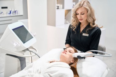 Avrupalı bir kadın güzellik uzmanı, güzellik salonundaki kanepede genç beyaz bir kadının IPL fotorejuvenation cihazıyla yüz derisini yüzüyor. Yüz bakımı ve gençleştirme kavramı