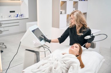 Avrupalı kadın kozmetik uzmanı, güzellik salonunda genç beyaz bir kadının yüz derisini mikronedle kaldırırken IPL fotorejuvenation cihazına basıyor. Cilt bakımı ve gençleştirme kavramı