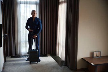 Ciddi bir Afro-Amerikan iş adamının elinde bavuluyla otel odasında gündüz kameraya bakması. İş gezisi, tatil ve seyahat kavramı