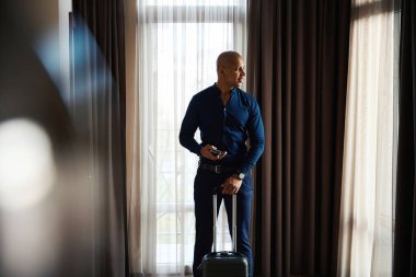 Gündüz vakti, elinde bavuluyla otel odasının camından içeri bakan Afro-Amerikan işadamının ön görüntüsü. İş gezisi, tatil ve seyahat kavramı