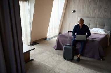 Odaklanmış Afro-Amerikan iş adamı. Gündüz vakti otel odasında yatarken dizüstü bilgisayar kullanıyor. İş gezisi, tatil ve seyahat kavramı