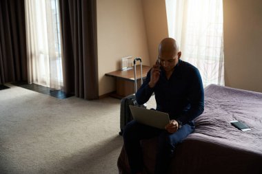 Gündüz vakti otel odasında yatarken cep telefonuyla konuşan ve dizüstü bilgisayar kullanan Afrikalı Amerikalı iş adamı. İş gezisi, tatil ve seyahat kavramı