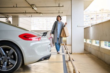 Otoparkta gülümseyen, alışveriş yapan, taşıyan, elinde kağıt torbalarla gezen mutlu kadın sürücü.