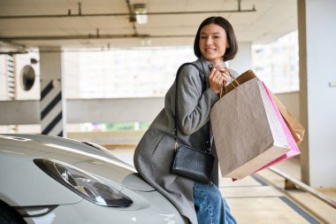 Günlük giysiler içinde güzel bir bayan alışveriş merkezinin otoparkında alışveriş yaparken mutlu ve kameraya bakarak gülümsüyor.