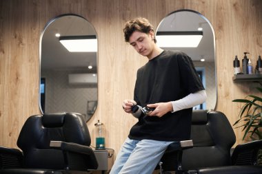 Yakışıklı berber sandalyeye yaslanıyor ve elinde saç makasıyla berber dükkanında fırça tutuyor.