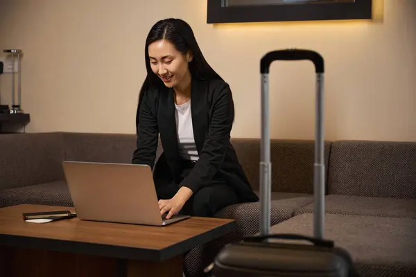 Stock image Smiling young lady sitting on couch and working on laptop from hotel room. Travel, business concept