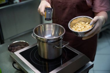 Kadın pastacı, buhar banyosundaki beyaz çikolata parçalarını eritmek için kaynamış suyun sıcaklığını ölçmek için termometre kullanıyor.