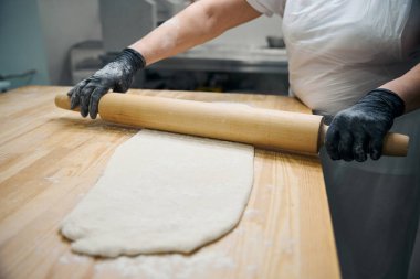 Tecrübeli aşçı hamurun bir kısmını oklavayla, üniformalı ve eldivenli bir kadını yuvarlıyor.