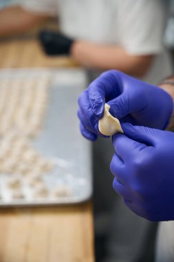 Koruyucu eldiven takan mutfak çalışanı küçük hamur tatlısı yapıyor. Masada bir sürü hazırlık var.