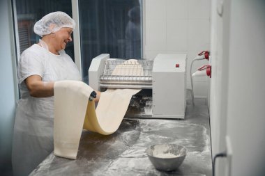 Deneyimli aşçı hamurun bir kısmını çarşaf makinesine sarar, atölyenin modern ekipmanları vardır.