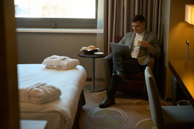 Elinde fincanla otel odasında çapraz bacaklı bir şekilde otururken dizüstü bilgisayara bakan ciddi bir iş adamı.