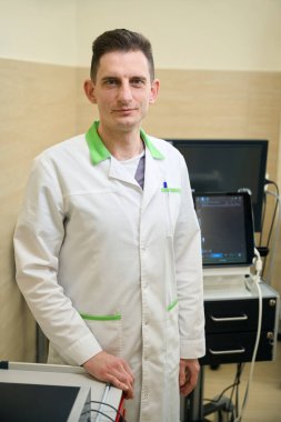 Tranquil doctor dressed in lab coat standing in examination room next to ultrasound machine clipart