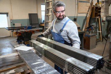 Foreman atölyede bir yığın alüminyum profili inceliyor, iş kıyafetleri içinde bir adam.